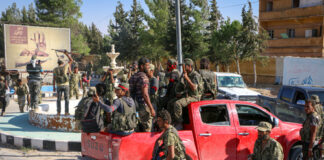 Group of soldiers in military gear gathered around a red pickup truck in an urban setting
