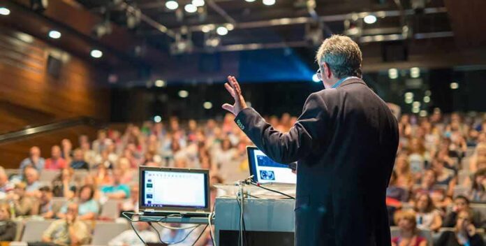 shutterstock_481869205 Speaker presenting to a large audience in an auditorium