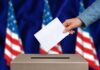 Hand inserting ballot into box, American flags background.