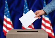 Hand inserting ballot into box, American flags background.